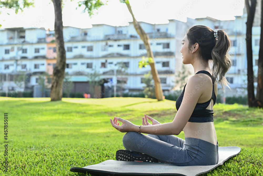 A woman is doing yoga in a park. The park is surrounded by buildings in the background. The woman is wearing a black top and gray pants.