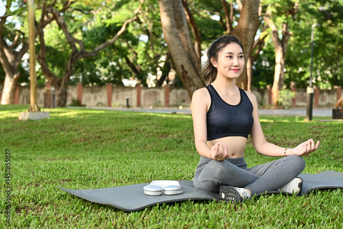 Obraz na płótnie A woman is sitting on a mat in a park, practicing yoga. She is wearing a black tank top and grey pants. The scene is peaceful and serene w salonie