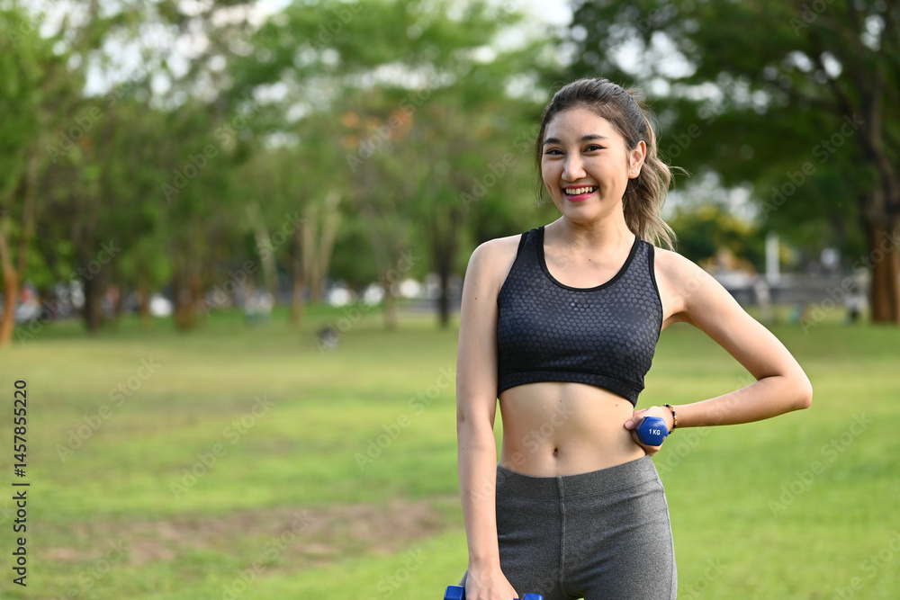 A woman is smiling and holding two dumbbells in a park. She is wearing a black tank top and gray pants.