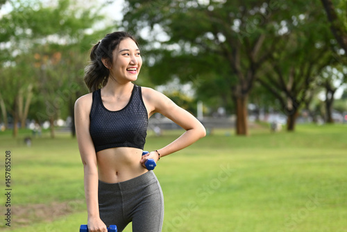 A woman is smiling and holding two dumbbells in a park. She is wearing a black tank top and gray pants.