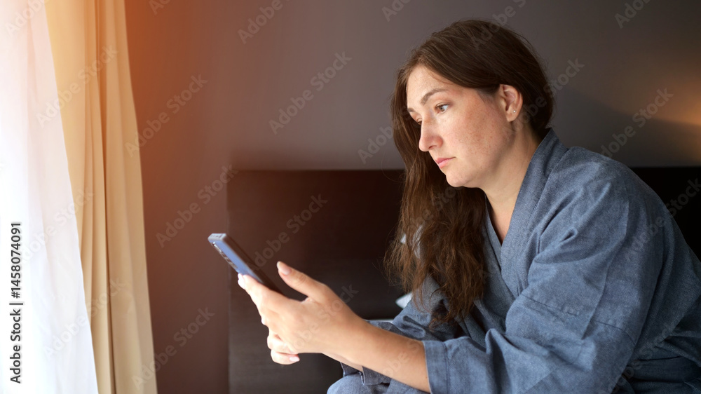 Female tourist sits on bed edge focused on phone in hotel apartment Female tourist sits on bed edge focused on phone in hotel apartment