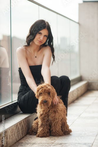 A fashionable young girl sits gracefully beside her fluffy dog in a modern urban setting. She smiles effortlessly while showcasing her trendy outfit, perfect for social media sharing