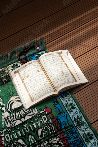Bandung, Indonesia - 02.11.2025:  Photo of opened Holy Quran with Arabic calligraphy and wooden beads or tasbih with natural daylight and prayer mat below it on wooden background
