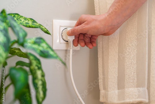 Close-up of hand inserting white electrical plug into wall socket next to houseplant, highlighting everyday electricity usage in domestic setting