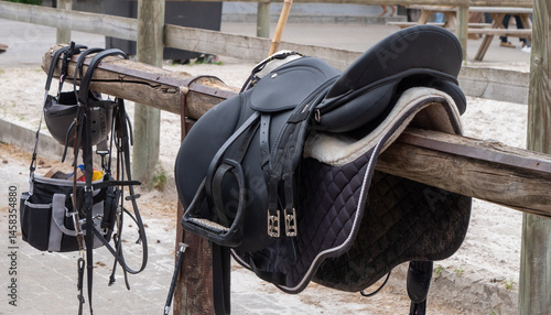 A black and white horse saddle is leaning against a wooden fence. The saddle is accompanied by a helmet and bridle.