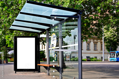 bus shelter at a busstop. blank billboard ad display. empty white lightbox sign. glass and aluminum frame structure. city transit station. bench inside. urban street setting. outdoor advertising