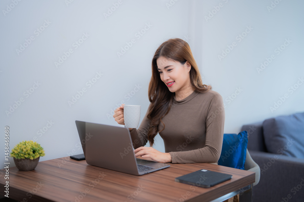 Fototapeta premium Young asian businesswoman drinking coffee while working on laptop sitting at her desk in home office, smart working online lifestyle and freelance job concept