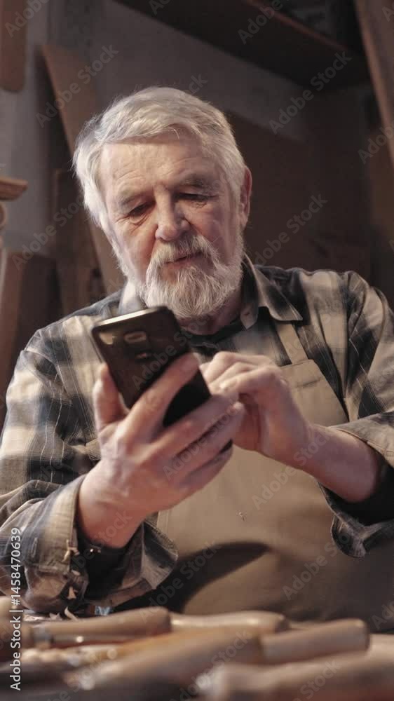 Elderly man with beard looking at mobile device in workshop. Focused on smartphone screen while holding phone with both hands. Wooden tools scattered on workbench around him. Tapping with fingers. Elderly man with beard looking at mobile device in workshop. Focused on smartphone screen while holding phone with both hands. Wooden tools scattered on workbench around him. Tapping with fingers.