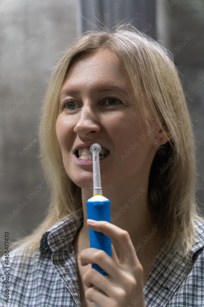 Young woman brushing her teeth with a modern electric toothbrush, maintaining good oral hygiene as part of her daily routine in a domestic bathroom setting. Vertical photo