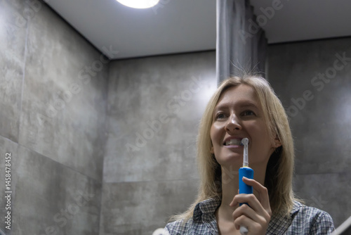 Obraz na płótnie Smiling woman brushing her teeth with an electric toothbrush in a modern bathroom, enjoying her daily oral hygiene routine and promoting dental health w salonie