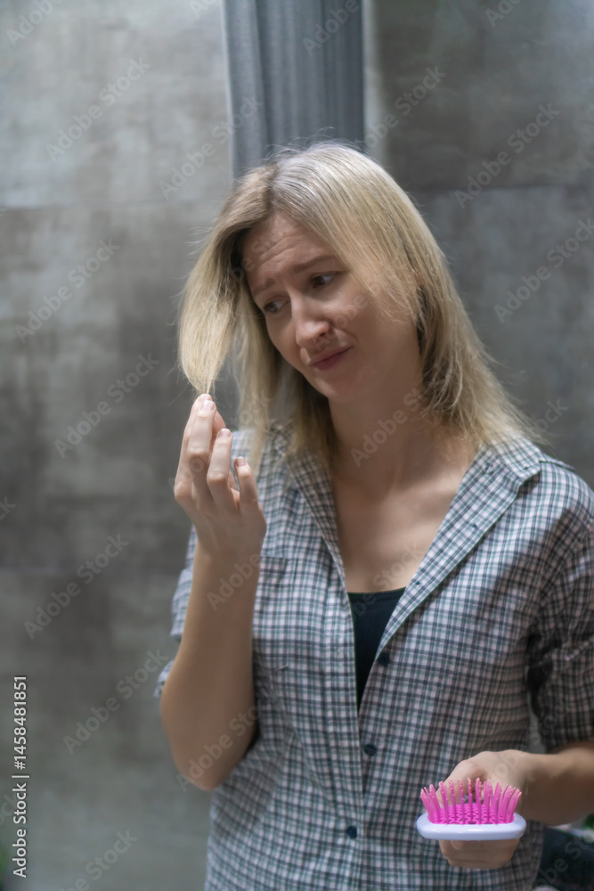 Young woman with blond hair wearing a plaid shirt, holding a pink brush and examining split ends or hair loss in the bathroom mirror, concerned about her hair health. Vertical photo