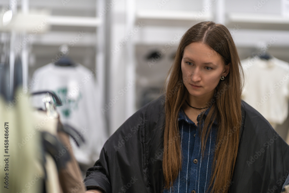 Young stylish woman choosing clothes, shopping in a fashion store, standing near clothes rack, examining garment, making purchasing decision