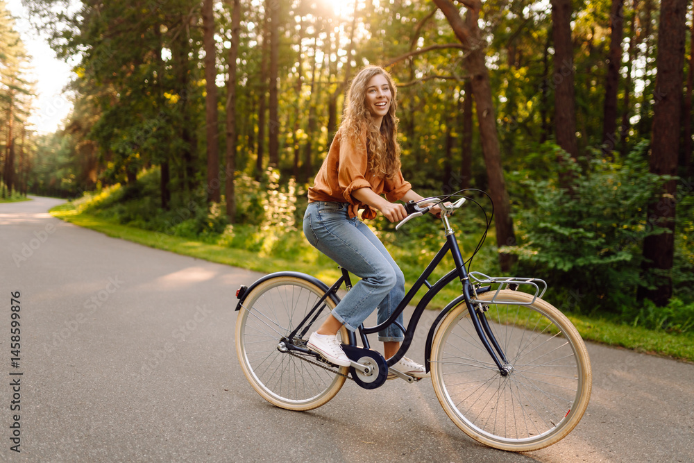 Fototapeta premium A joyful woman with curly hair rides a bicycle along a path in a sunny park. Young woman having fun and enjoying a day off outdoors. Active lifestyle.