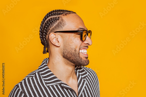 Side view portrait of a cheerful young multiracial man with braids wearing glasses, smiling and showing teeth against a vibrant yellow background.