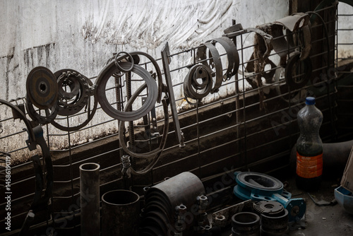 Various greasy metal parts hang from a wire mesh against a dirty window, with more tools and components scattered on a workbench below. A cardboard box sits to the left.