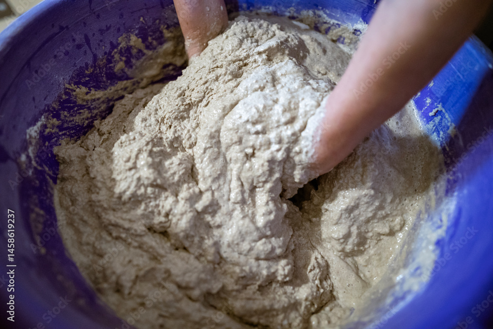 Hands kneading dough in a large plastic container during the process of preparing homemade bread. The dough is sticky and textured, showing active mixing.