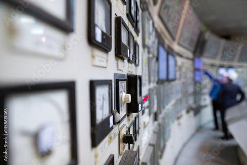 Obraz na płótnie Control room featuring control panels with multiple dials, switches, and meters. Two workers in hard hats stand in the background, with one pointing at a screen, overseeing industrial operations. w salonie