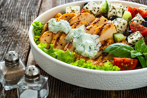 Foto Grilled sliced chicken breast with tzatziki sauce and Greek salad on wooden tabl