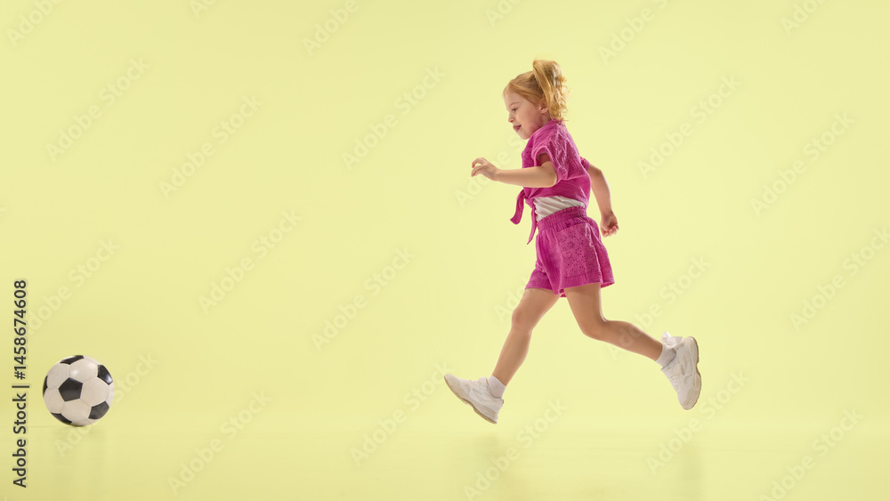 Joyful child girl in pink shorts and shirts running towards soccer ball, playing against yellow background. Concept of childhood, summer vacation, active lifestyle, fun, sport. Banner