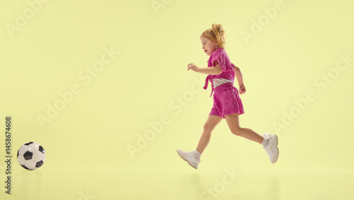 Obraz na płótnie Joyful child girl in pink shorts and shirts running towards soccer ball, playing against yellow background. Concept of childhood, summer vacation, active lifestyle, fun, sport. Banner w salonie