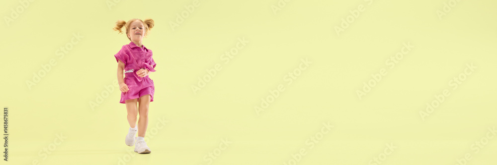 Low angle view of happy child girl with ponytail hairstyle, walking in pink outfit, smiling playfully against yellow background. Concept of childhood, summer vacation, active lifestyle, fun, leisure