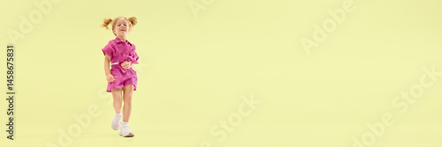 Obraz na płótnie Low angle view of happy child girl with ponytail hairstyle, walking in pink outfit, smiling playfully against yellow background. Concept of childhood, summer vacation, active lifestyle, fun, leisure w salonie