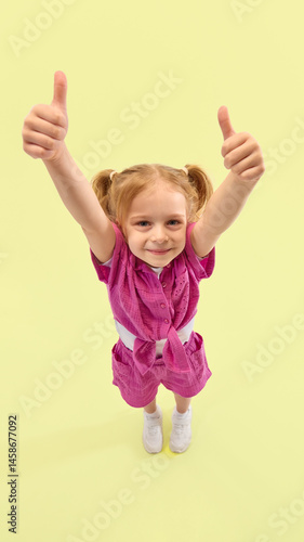Cheerful girl lifts both thumbs up, smiling directly in camera, showing positive mood and happiness against yellow background. Concept of childhood, summer vacation, active lifestyle, fun