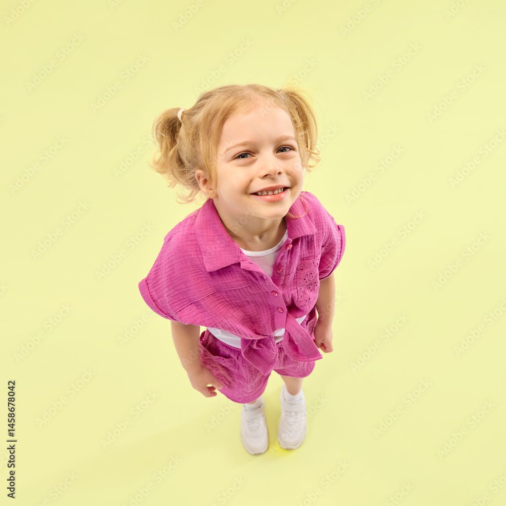 Smiling girl with ponytails wearing pink shirt and shorts, poses joyfully against yellow background. Happiness and leisure. Concept of childhood, summer vacation, active lifestyle, fun
