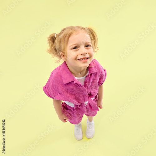 Obraz na płótnie Smiling girl with ponytails wearing pink shirt and shorts, poses joyfully against yellow background. Happiness and leisure. Concept of childhood, summer vacation, active lifestyle, fun w salonie