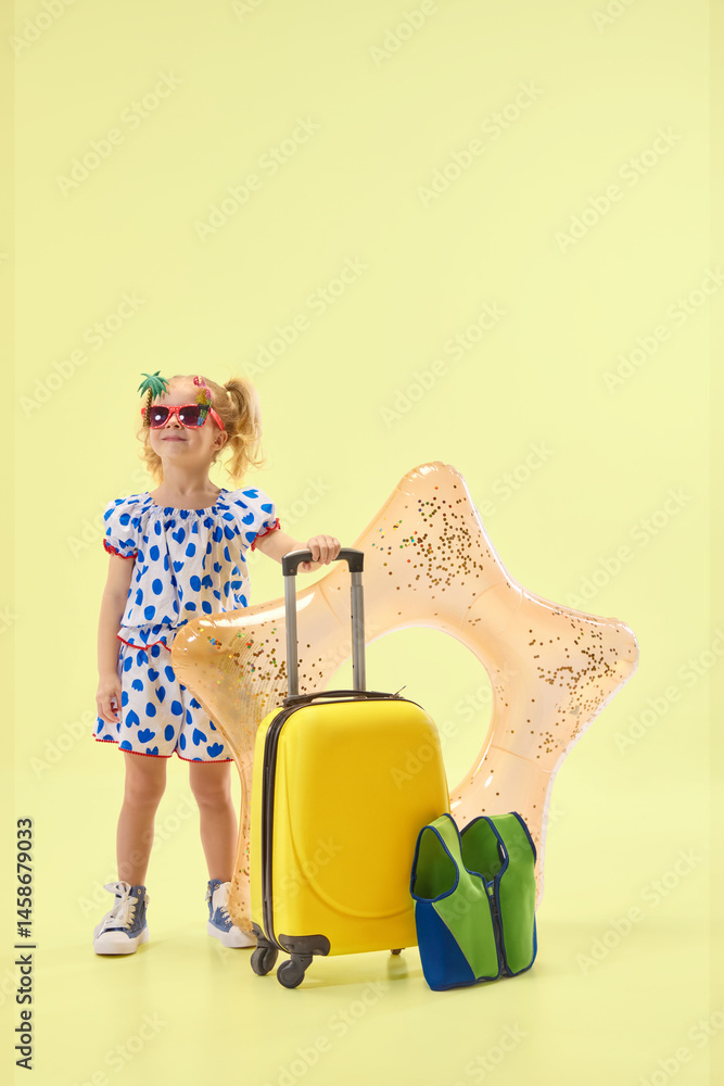 Happy little girl in blue dress and sunglasses standing with float ring and packed suitcase against yellow background. Concept of childhood, summer vacation, travelling, trip
