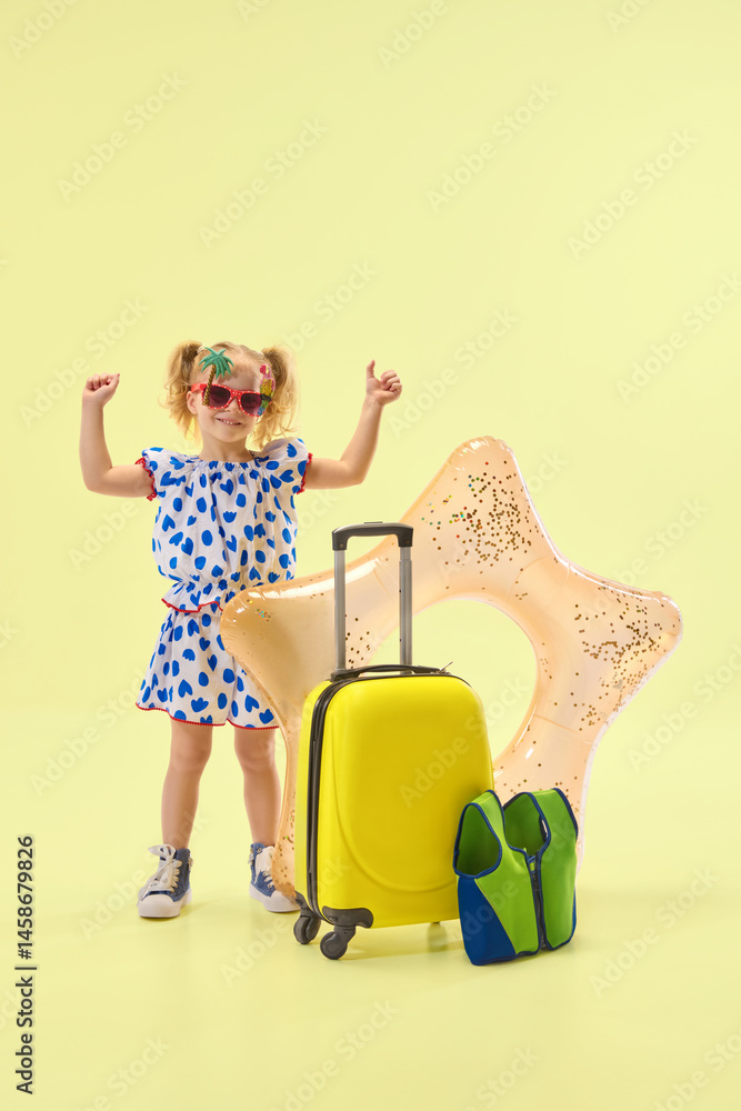 Cheerfully girl raising hands in excitement, standing near suitcase and inflatable float, expressing happiness against yellow background. Concept of childhood, summer vacation, travelling, trip