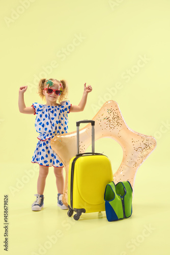 Obraz na płótnie Cheerfully girl raising hands in excitement, standing near suitcase and inflatable float, expressing happiness against yellow background. Concept of childhood, summer vacation, travelling, trip w salonie