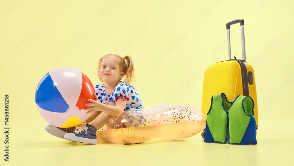 Happy girl in blue dress sitting on float with beach ball beside suitcase, expressing happiness and joy against yellow background. Concept of childhood, summer vacation, travelling, trip