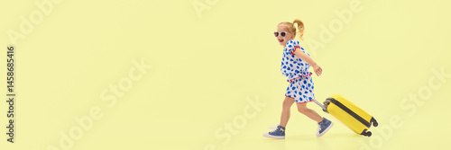 Obraz na płótnie Joyful girl drags yellow suitcase while walking confidently against yellow background, expressing energy and excitement. Concept of childhood, summer trip, joy, activity, motion, adventure. w salonie