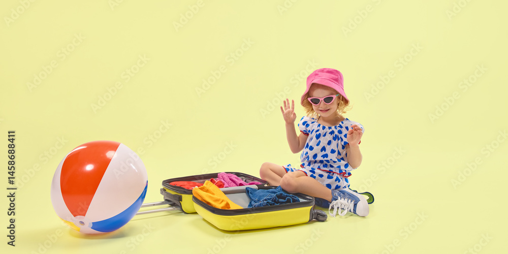 Little girl waves hand sitting near suitcase filled with colorful clothes with beach ball on floor, smiling during holiday preparation on yellow background. Concept of childhood, summer, vacation