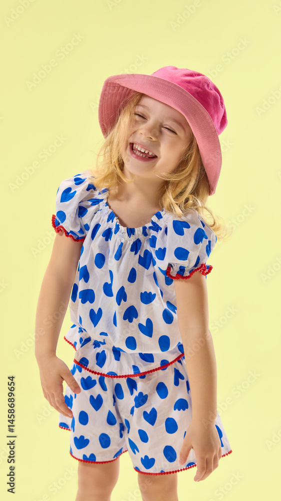 Smiling girl in pink hat laughs joyfully, dressed in cute dress, showing happy mood and carefree spirit against yellow background. Concept of childhood, summer, vacation, play, activity, fun.