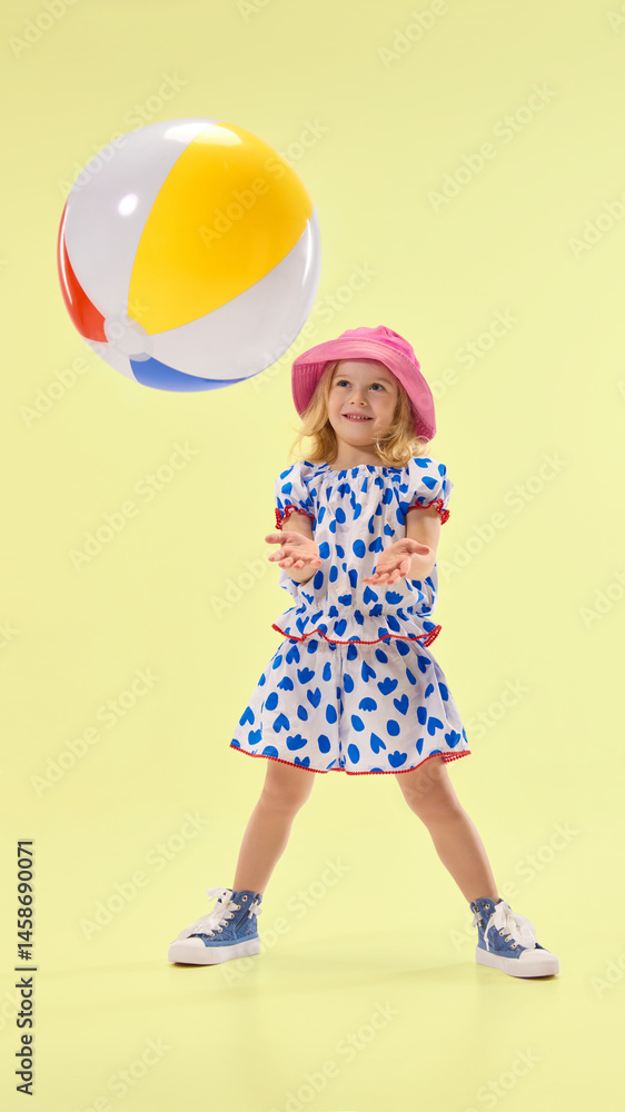 Happy girl plays with beach ball, catching it with open arms, showing excitement against vibrant yellow backdrop. Concept of childhood, summer, vacation, play, activity, fun.