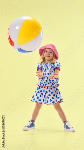 Obraz na płótnie Happy girl plays with beach ball, catching it with open arms, showing excitement against vibrant yellow backdrop. Concept of childhood, summer, vacation, play, activity, fun. w salonie