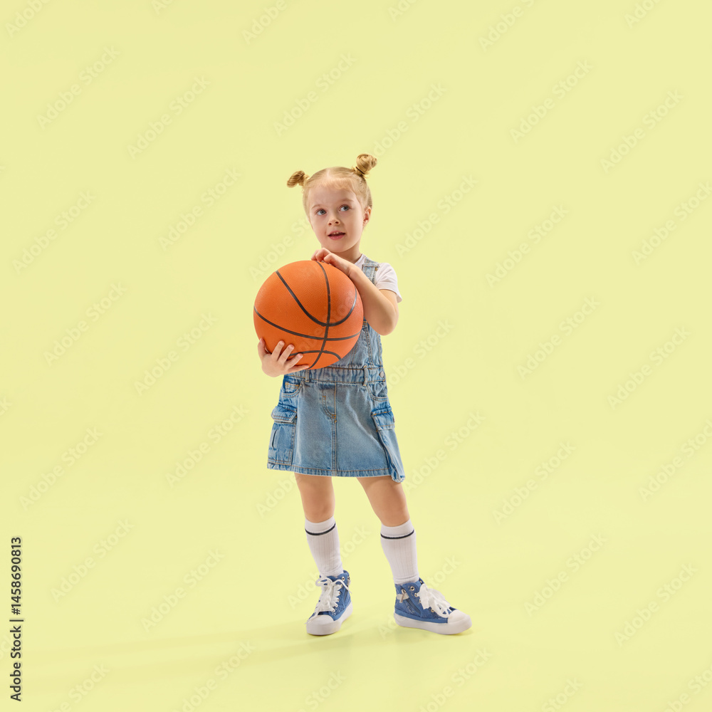 Young girl holds basketball, wearing denim dress and white socks, looking curious and calm, posing against yellow backdrop. Concept of childhood, summer, vacation, play, activity, fun.