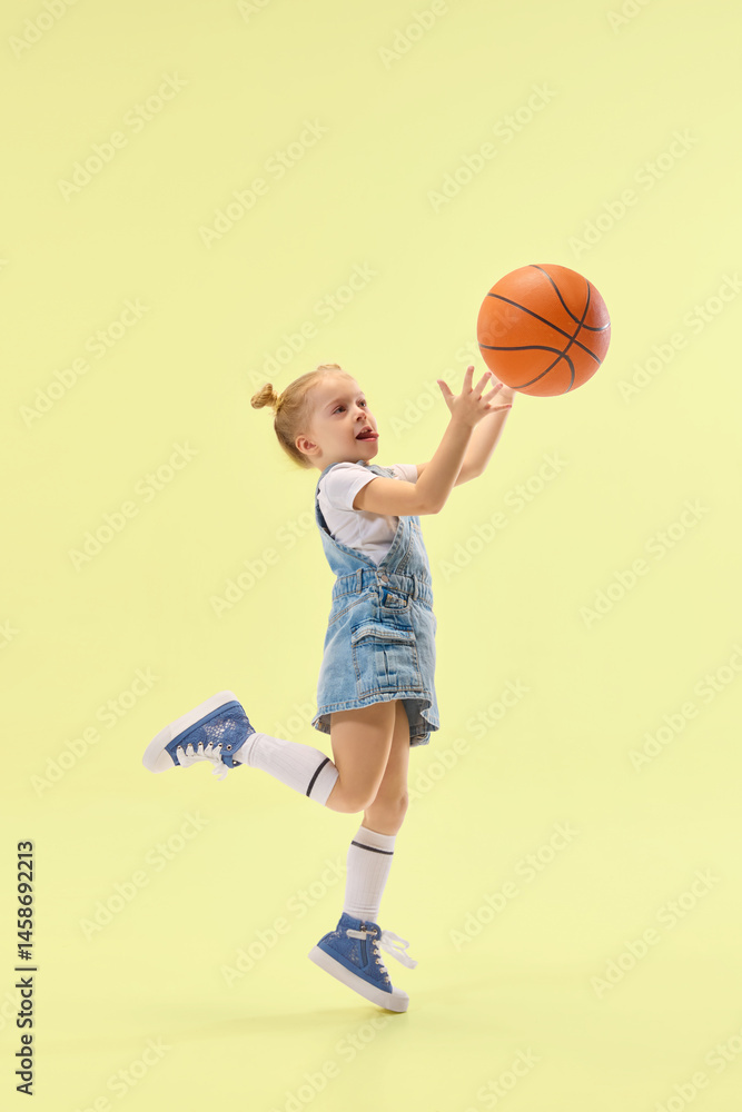 Active girl jumps, catching basketball midair, showing sporty action with energetic motion, wearing denim jumper over yellow pastel backdrop. Concept of childhood, summer, vacation, play, activity