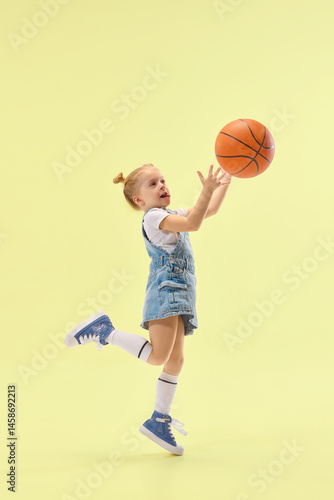 Obraz na płótnie Active girl jumps, catching basketball midair, showing sporty action with energetic motion, wearing denim jumper over yellow pastel backdrop. Concept of childhood, summer, vacation, play, activity w salonie