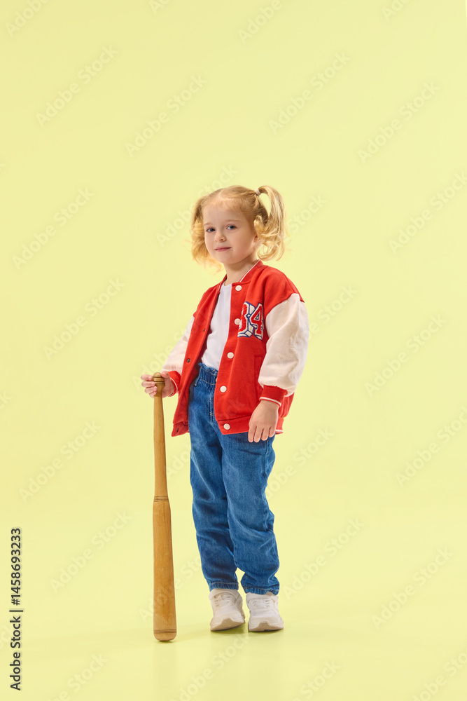 Smiling girl in red varsity jacket holds wooden baseball bat, ready to play against yellow background. Concept of childhood, sport, activity, game, fun, leisure