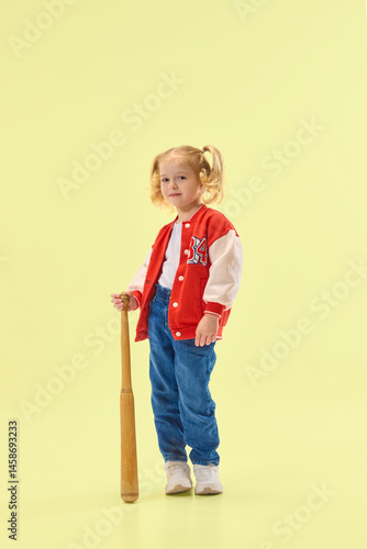 Obraz na płótnie Smiling girl in red varsity jacket holds wooden baseball bat, ready to play against yellow background. Concept of childhood, sport, activity, game, fun, leisure w salonie