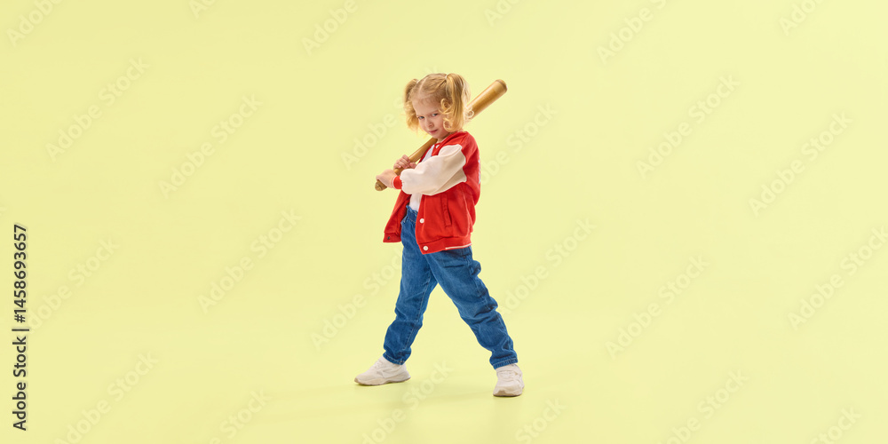 Smiling girl in red varsity jacket holds wooden baseball bat over shoulder, confidently standing against yellow background. Concept of childhood, sport, activity, game, fun, leisure