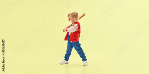 Obraz na płótnie Smiling girl in red varsity jacket holds wooden baseball bat over shoulder, confidently standing against yellow background. Concept of childhood, sport, activity, game, fun, leisure w salonie