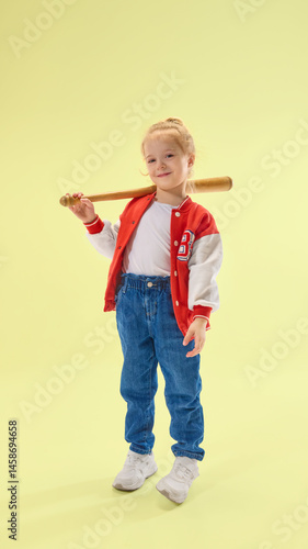Obraz na płótnie Cheerful girl holds bat on shoulder, smiling confidently, in red jacket and denim pants over yellow background. Concept of childhood, sport, activity, game, fun, leisure w salonie