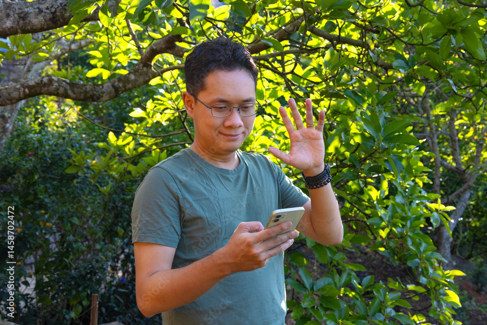 An Asian man is using his phone, texting or video calling in a garden An Asian man is using his phone, texting or video calling in a garden
