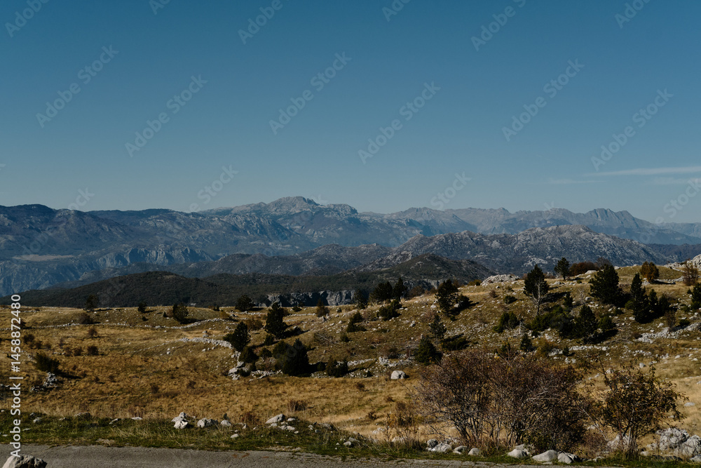 Fototapeta premium Scenic view of the Montenegro mountains under a clear blue sky. Rolling hills with sparse trees and rocky terrain stretch toward the majestic mountain range in the distance.