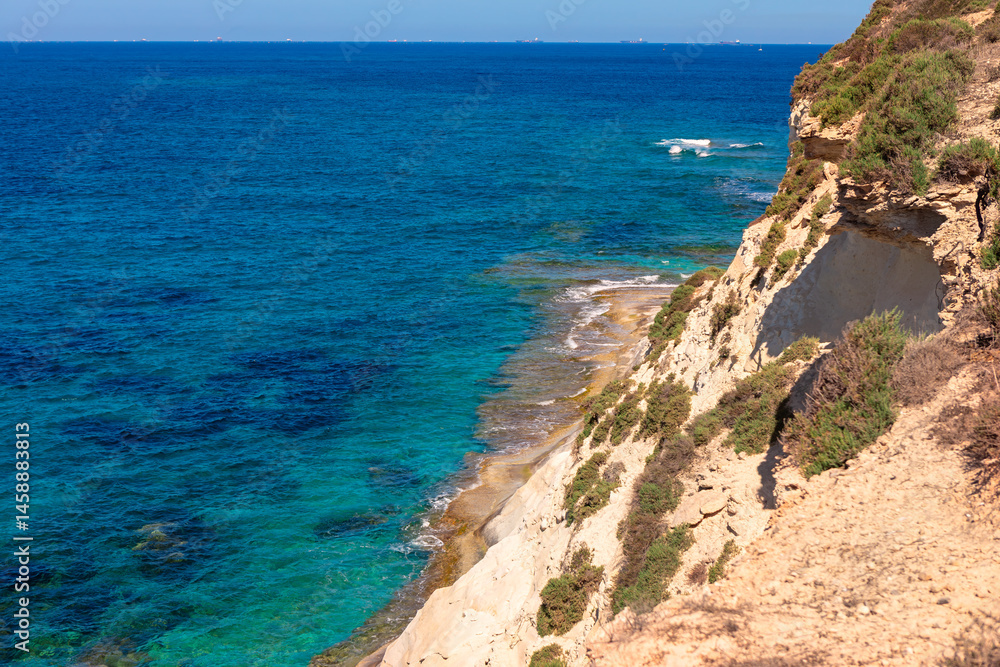 Stunning coastal scene with clear turquoise waters gently lapping against a rocky shoreline and steep, vegetation covered cliffs. Deep blue ocean stretches to horizon under a bright, cloudless sky