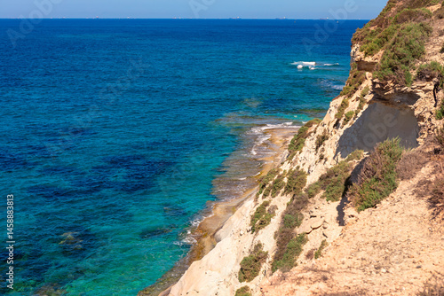 Obraz na płótnie Stunning coastal scene with clear turquoise waters gently lapping against a rocky shoreline and steep, vegetation covered cliffs. Deep blue ocean stretches to horizon under a bright, cloudless sky w salonie
